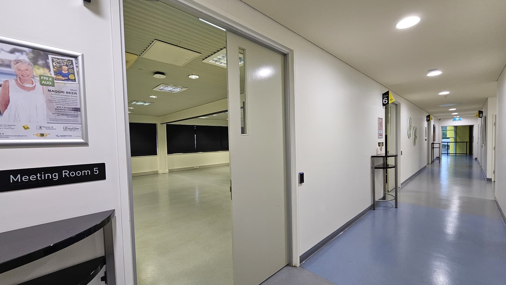 On the left is an open door with a sign reading "Meeting Room 5" and a poster above it. Inside the room, black curtains cover the windows, and a light grey floor. On the right, there is a corridor with a small table holding a decorative item and a sign indicating "6". The corridor has a light blue and grey floor and white walls. In the background, the corridor continues with more doors and a bright light at the end. The ceiling is white with recessed lighting and ventilation panels.