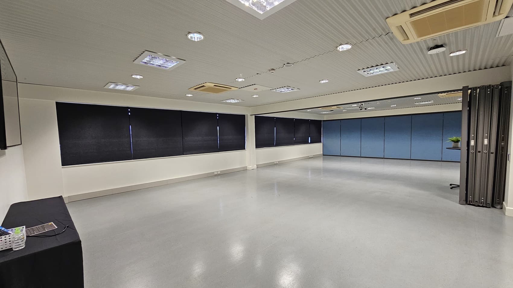On the left, there is a black table with a basket containing various items. On the right, there is a partition wall with a small table holding a green plant. In the middle, there is a large open space with light grey flooring. The background features black curtains covering windows and a blue partition wall. The ceiling is white with multiple recessed lights and air conditioning units.