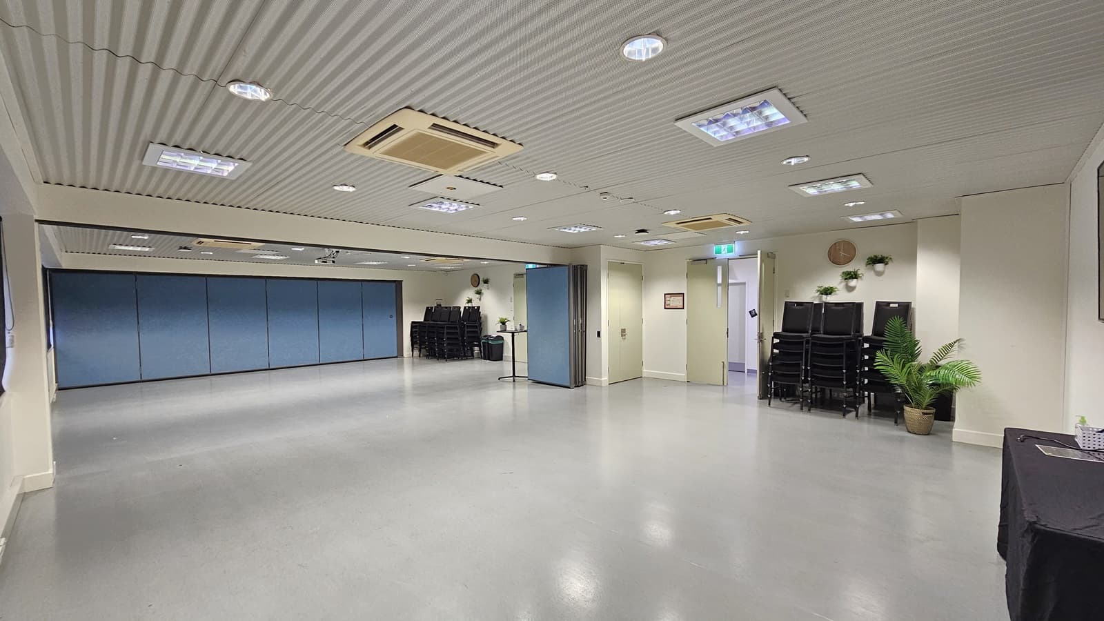 On the left, there is a large open space with blue partition walls. On the right, there is a stack of black chairs next to a potted plant and a small table with a water dispenser. In the middle, there is a column dividing the space. The background features more stacked chairs and a blue partition wall. The floor is light grey, and the ceiling is white with recessed lighting and air conditioning units.
