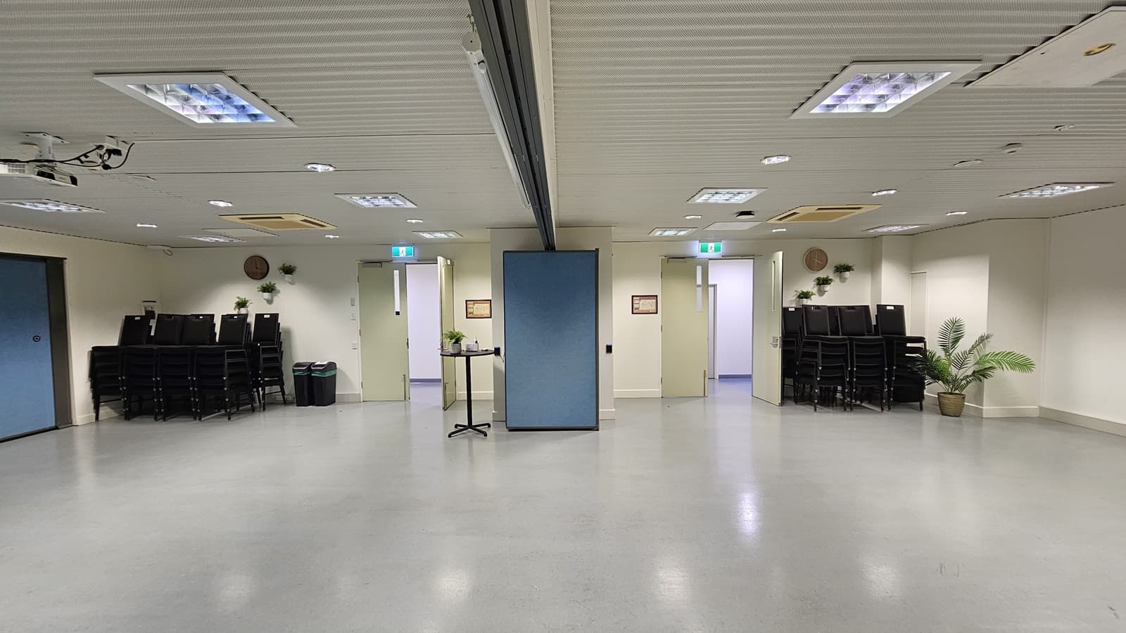 On the left, there is a stack of black chairs next to a black bin and a door with an exit sign above it. On the right, there is another stack of black chairs next to a potted plant and a door with an exit sign above it. In the middle, there is a blue partition wall with a small table holding a plant and a sign on the wall. The floor is light grey, and the ceiling is white with recessed lighting and air conditioning units.