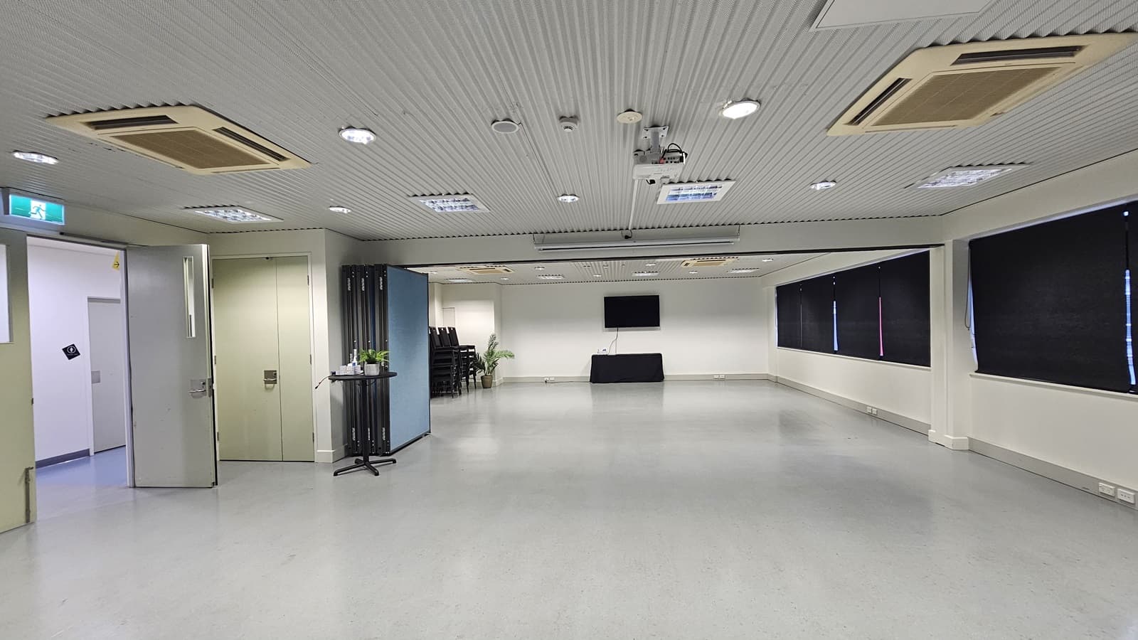 On the left, there is a double door with an exit sign above it and a small table with a plant. On the right, there are black curtains covering the windows. In the middle, there is a large open space with a black table against the far wall and a wall-mounted television above it. The background features a stack of black chairs near the back wall. The floor is light grey, and the ceiling is white with recessed lighting and multiple air conditioning units.