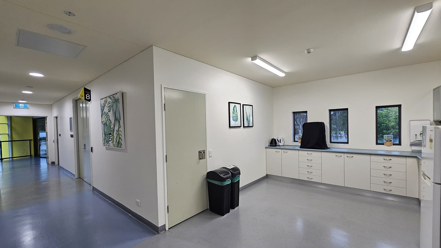 On the left, there is a corridor with white walls, a blue floor, and several doors, one with a sign labelled "8". On the right, there is a kitchenette area with white cabinets, a countertop, a kettle, and three small windows. In the middle, there is a white door next to two black bins with green lids. The background features a yellow door at the end of the corridor. The ceiling is white with recessed lighting and ventilation panels.