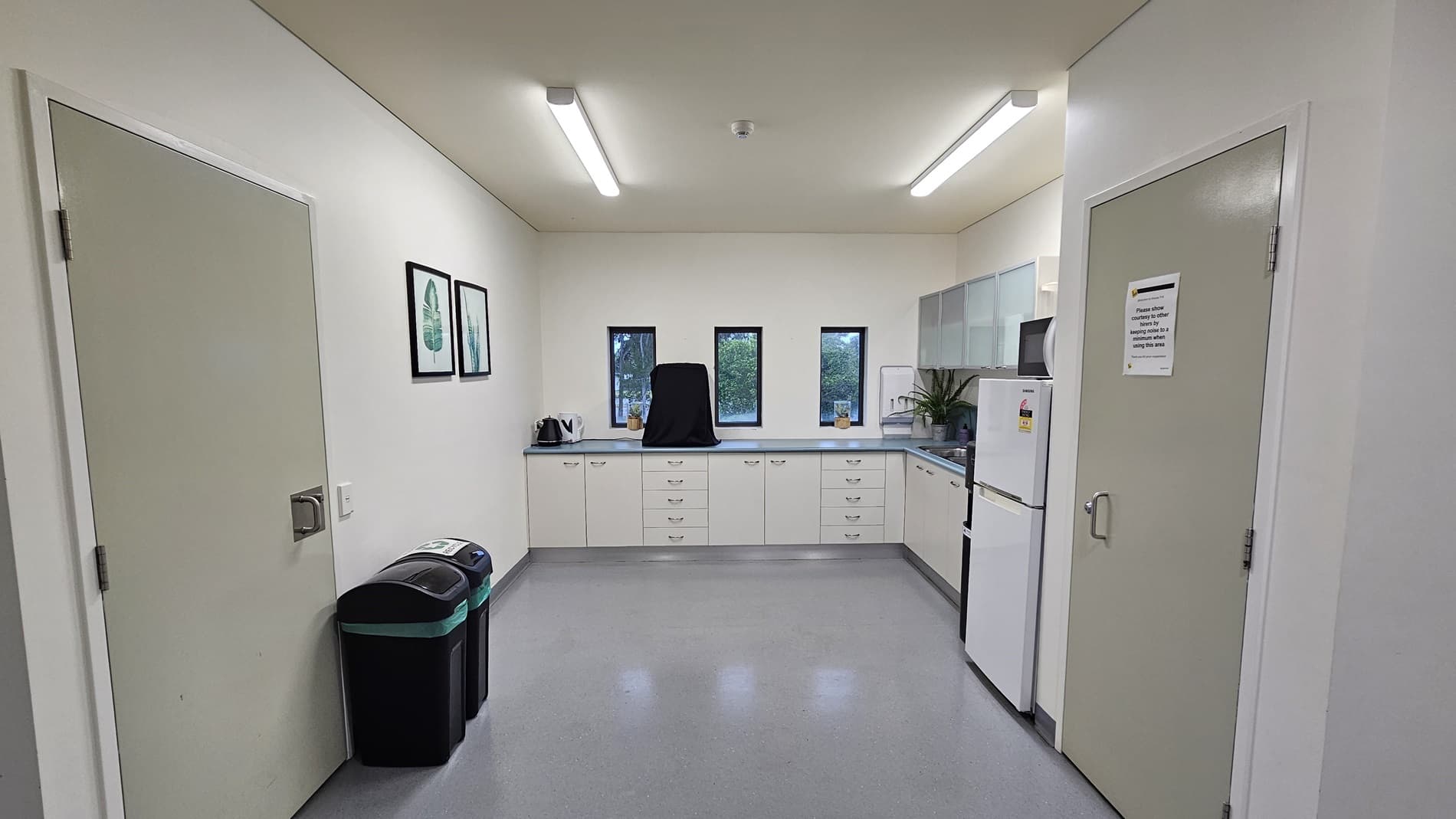 On the left, there is a white door next to two black bins with green lids. On the right, there is another white door with a sign on it and a white refrigerator beside it. In the middle, there is a kitchenette with white cabinets, a countertop, a kettle, and three small windows above the counter. The floor is light grey, and the ceiling is white with recessed lighting.