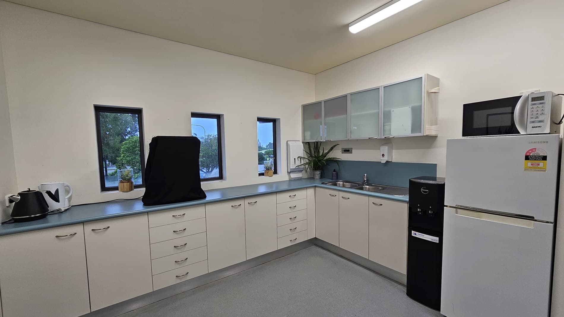 On the left, there is a white kettle and a small potted plant on a blue countertop. On the right, there is a white refrigerator with a microwave on top and a black water dispenser beside it. In the middle, there are three small windows above the countertop, a sink with a plant next to it, and white cabinets with frosted glass doors. The floor is light grey, and the ceiling is white with a long fluorescent light fixture.