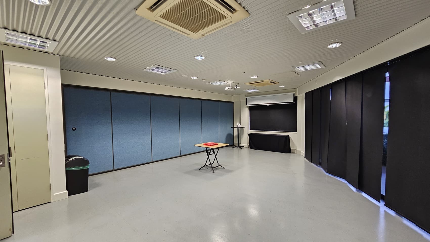 On the left, there is a white door next to a black bin with a green lid. On the right, there are black curtains covering the windows. In the middle, there is a small table with a red object on it. The background features blue partition walls and a black table with a whiteboard above it. The floor is light grey, and the ceiling is white with recessed lighting and an air conditioning unit.