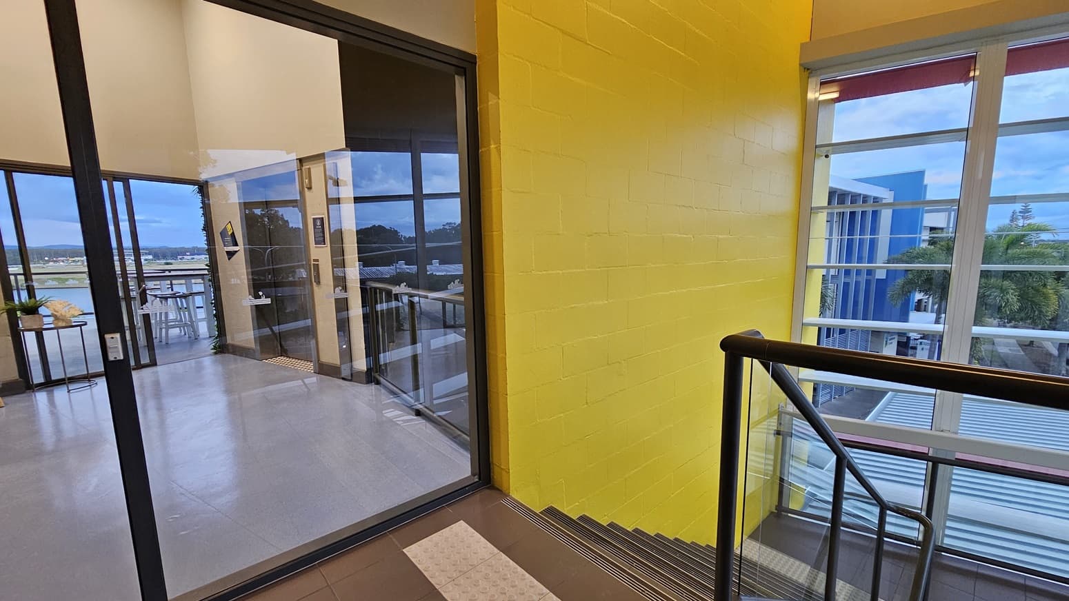 On the left, there is a glass door leading to a room with grey flooring, white tables, and chairs. On the right, there is a yellow brick wall next to a staircase with metal railings. In the background, large windows reveal a view of blue buildings and palm trees. The floor in the foreground is dark grey, and the ceiling is white with recessed lighting.