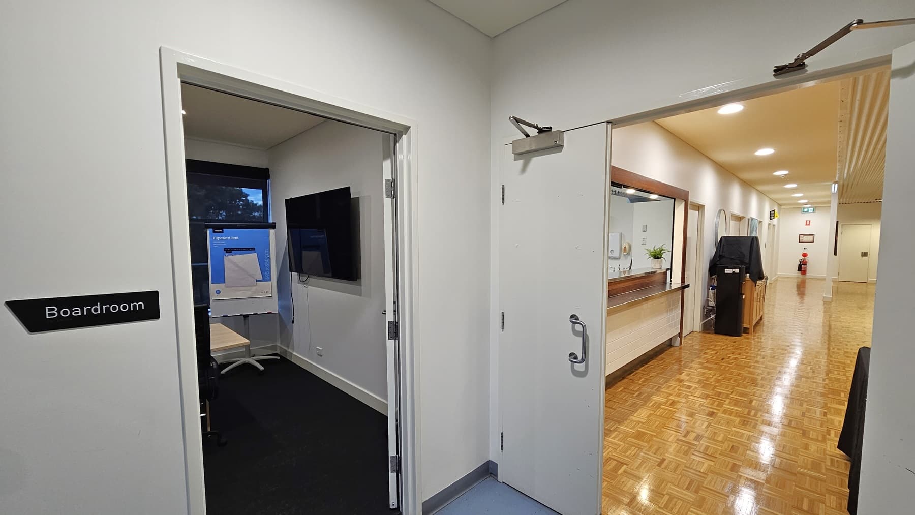 On the left, there is an open door with a sign labelled "Boardroom," revealing a room with a black carpet, a wall-mounted television, and a flip chart. On the right, there is a corridor with a wooden parquet floor, white walls, and a reception desk with a mirror above it. In the background, the corridor extends to a fire extinguisher and an exit sign. The ceiling is white with recessed lighting.