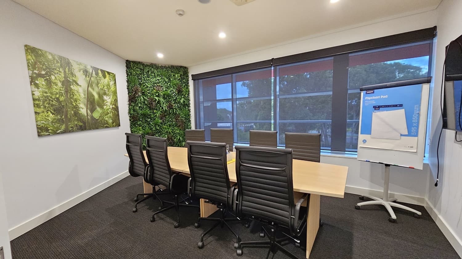 On the left, there is a large framed picture of a forest scene on a white wall and a section of wall covered in green foliage. On the right, there are large windows with roller blinds partially down and a flip chart on a stand. In the middle, there is a long wooden conference table surrounded by black office chairs. The floor is covered in dark grey carpet, and the ceiling is white with recessed lighting.