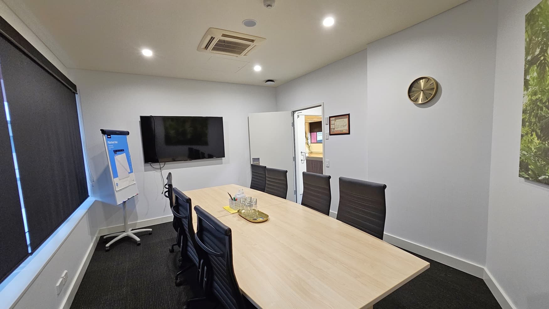 On the left, there is a flip chart on a stand next to a large wall-mounted television. On the right, there is a clock on the wall and a framed certificate above an open door leading to another room. In the middle, there is a long wooden conference table with black office chairs around it, and a tray with glasses and a water jug on the table. The floor is covered in dark grey carpet, and the ceiling is white with recessed lighting and an air conditioning unit.