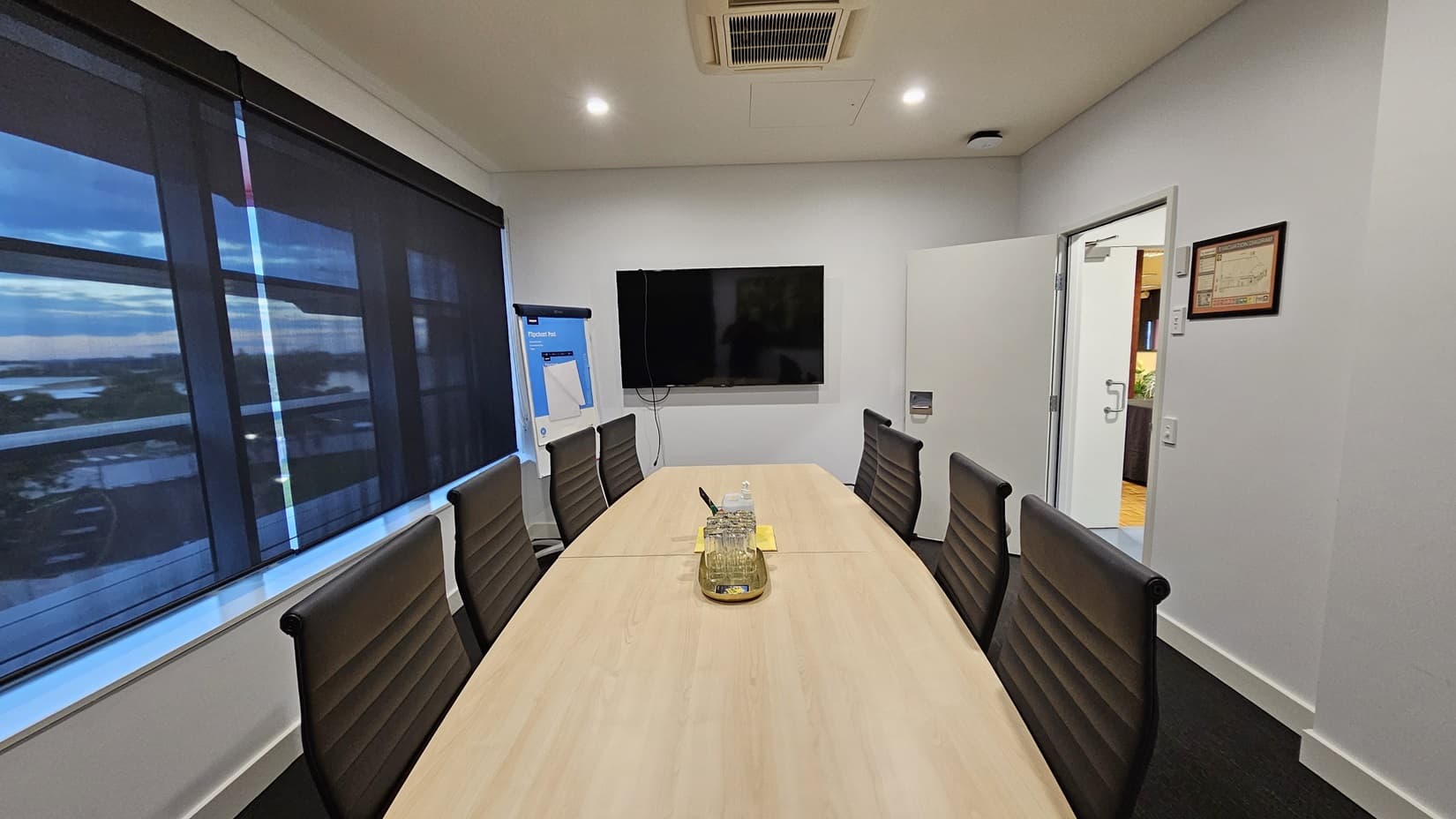 On the left, there are large windows with black roller blinds partially down and a flip chart on a stand. On the right, there is an open door leading to another room and a framed certificate on the wall. In the middle, there is a long wooden conference table with black office chairs around it, and a tray with a water jug and glasses on the table. The floor is covered in dark grey carpet, and the ceiling is white with recessed lighting and an air conditioning unit.