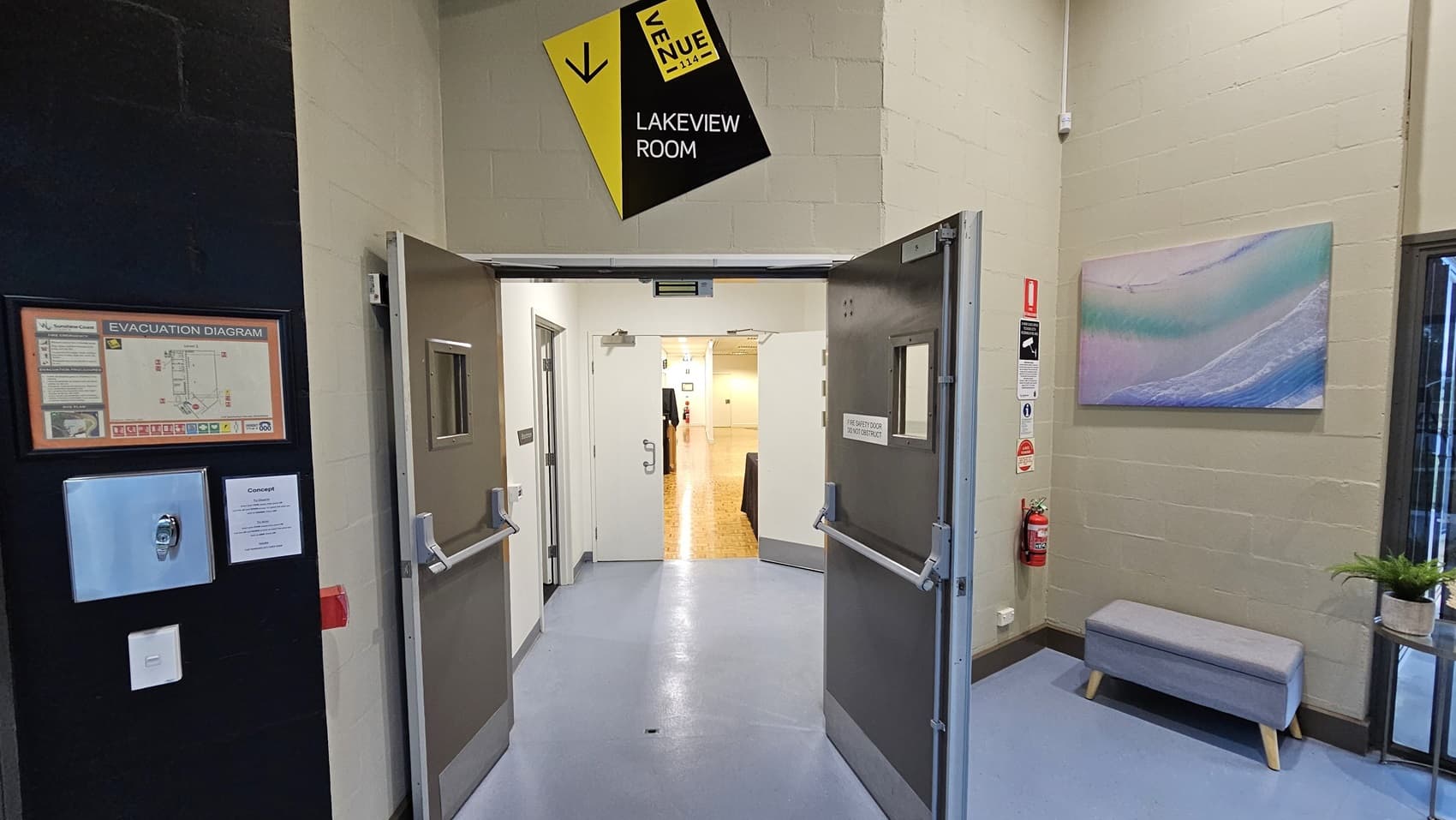 On the left, there is a wall-mounted evacuation diagram and a hand sanitiser dispenser. On the right, there is a framed picture of a beach scene above a grey bench with a potted plant next to it. In the middle, double doors open to a corridor with a wooden parquet floor and white walls. The floor in the foreground is light grey, and the ceiling is white with recessed lighting. A yellow sign above the doors reads "Lakeview Room."