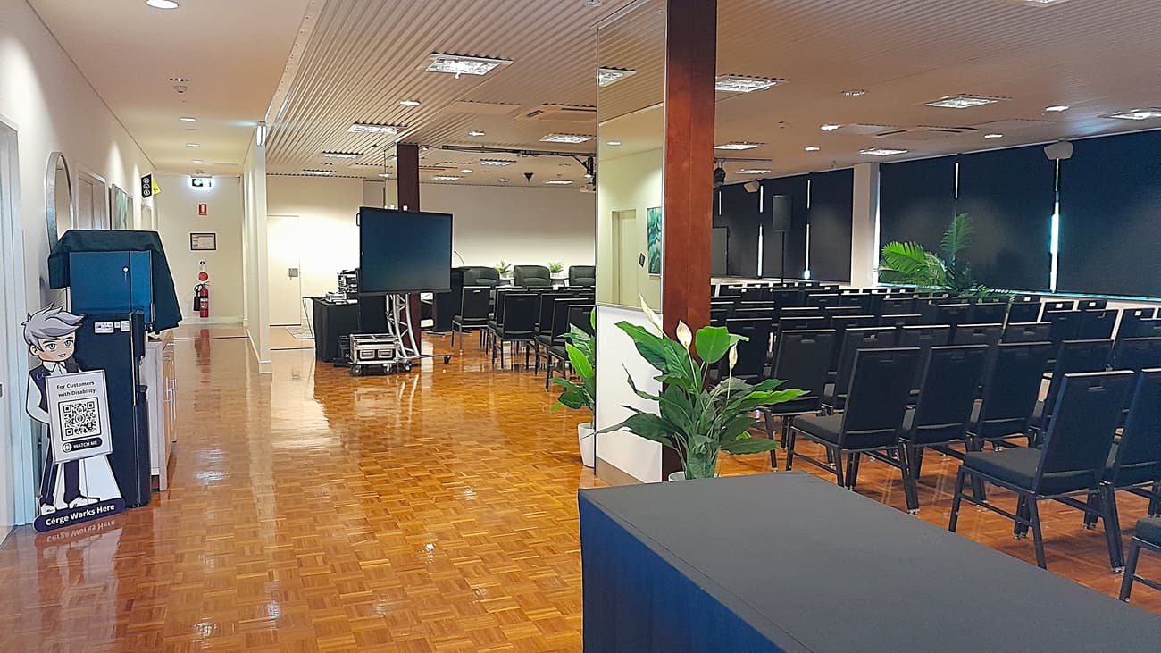 On the left, there is a corridor with a wooden parquet floor, white walls, and a blue recycling bin with a cartoon character on it. On the right, there are rows of black chairs facing a large screen, with black roller blinds partially down on the windows. In the middle, there is a large television on a stand and a potted plant near a wooden pillar. The ceiling is white with recessed lighting and ventilation panels.