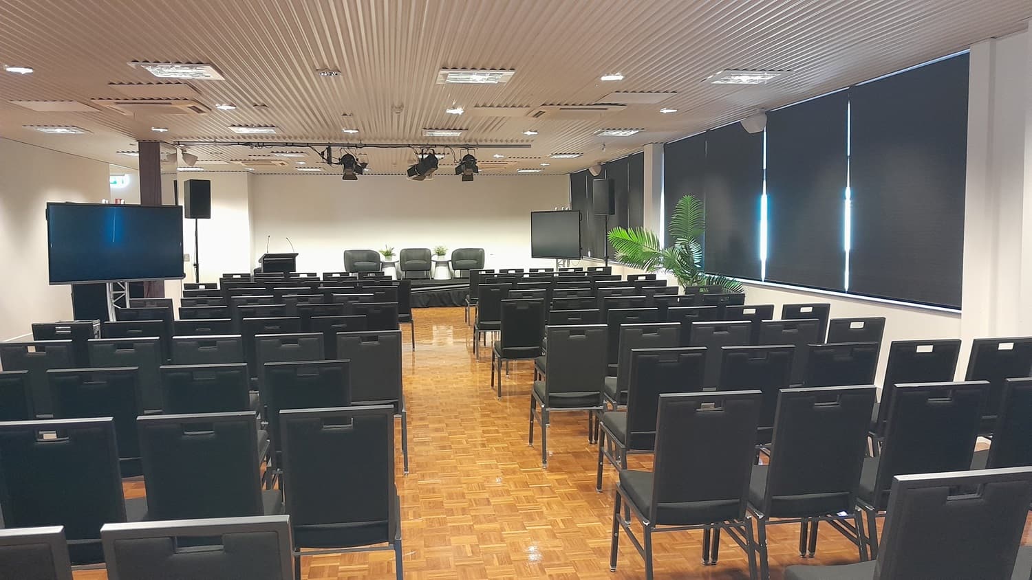 On the left, there is a large television on a stand and a black speaker. On the right, there are large windows with black roller blinds partially down and a potted plant near the corner. In the middle, there are rows of black chairs facing a stage with a podium, three black chairs, and a small table. The floor is covered in wooden parquet, and the ceiling is white with recessed lighting and ventilation panels.