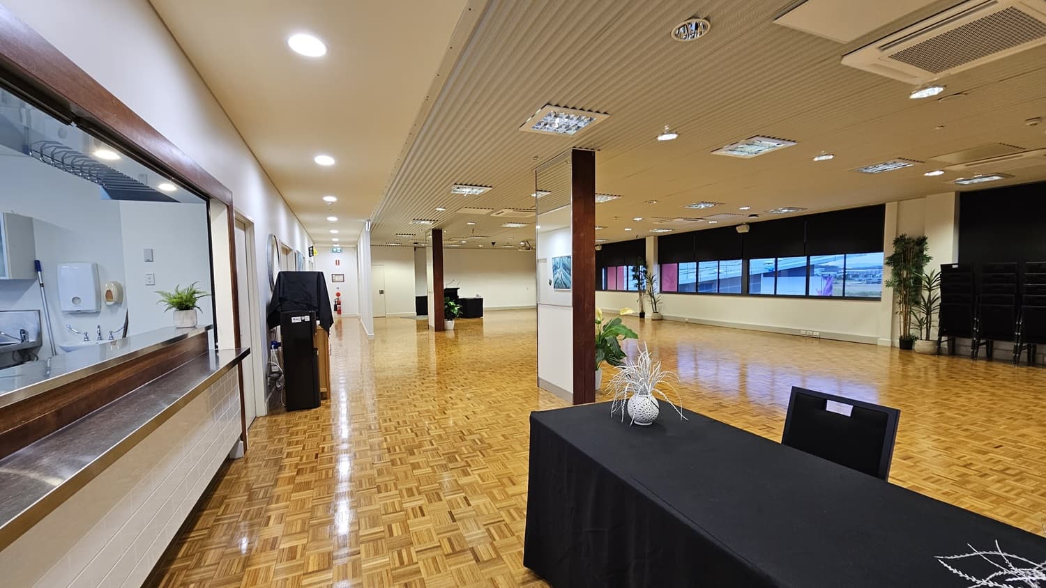 On the left, there is a long counter with a service window, a potted plant, and a black cabinet. On the right, there are large windows with black roller blinds partially down and a stack of black chairs in the corner. In the middle, there is a black table with a decorative vase and a single black chair. The floor is covered in wooden parquet, and the ceiling is white with recessed lighting and ventilation panels.