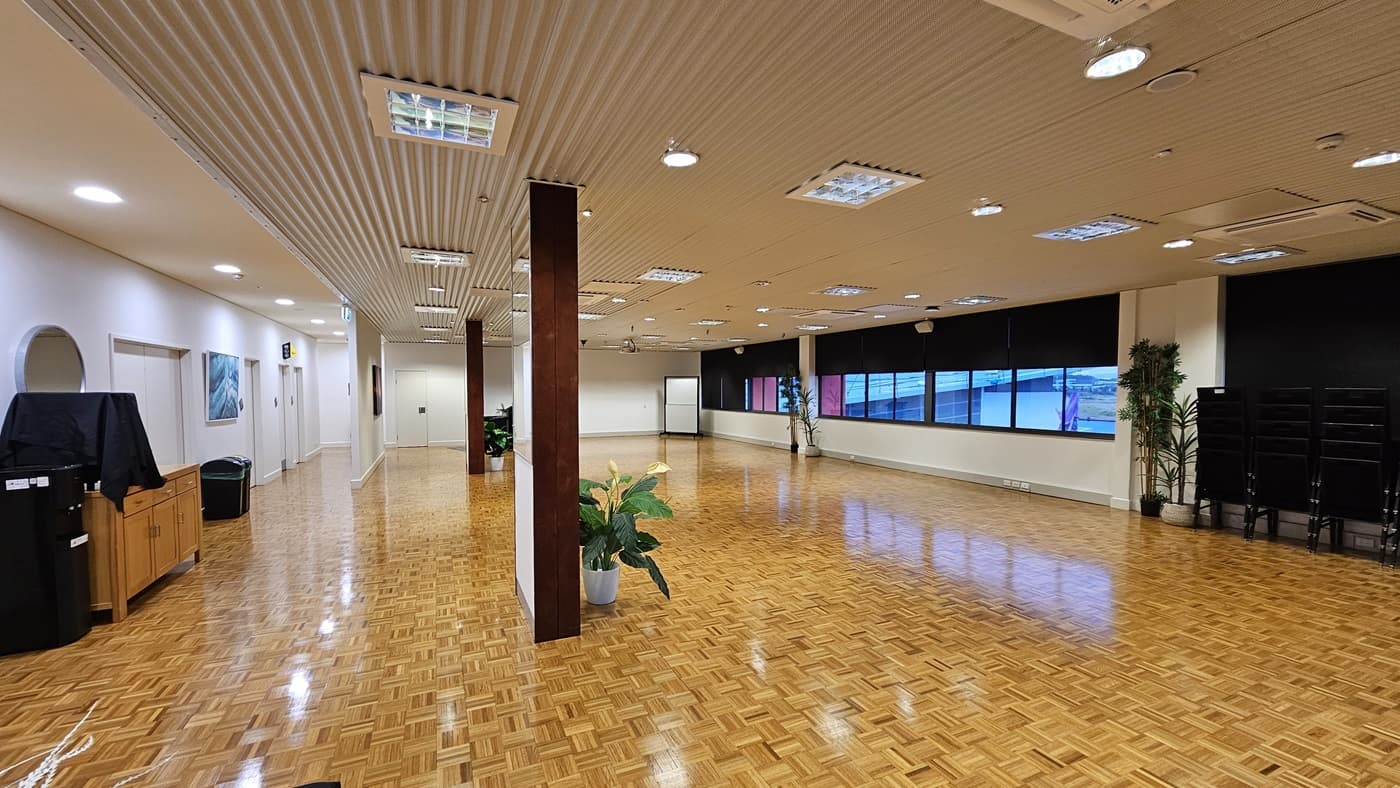 On the left, there is a wooden cabinet with a black cover, a potted plant, and a black bin. On the right, there are large windows with black roller blinds partially down, a stack of black chairs, and two potted plants. In the middle, there is a wooden pillar with a potted plant at its base. The floor is covered in wooden parquet, and the ceiling is white with recessed lighting and ventilation panels.