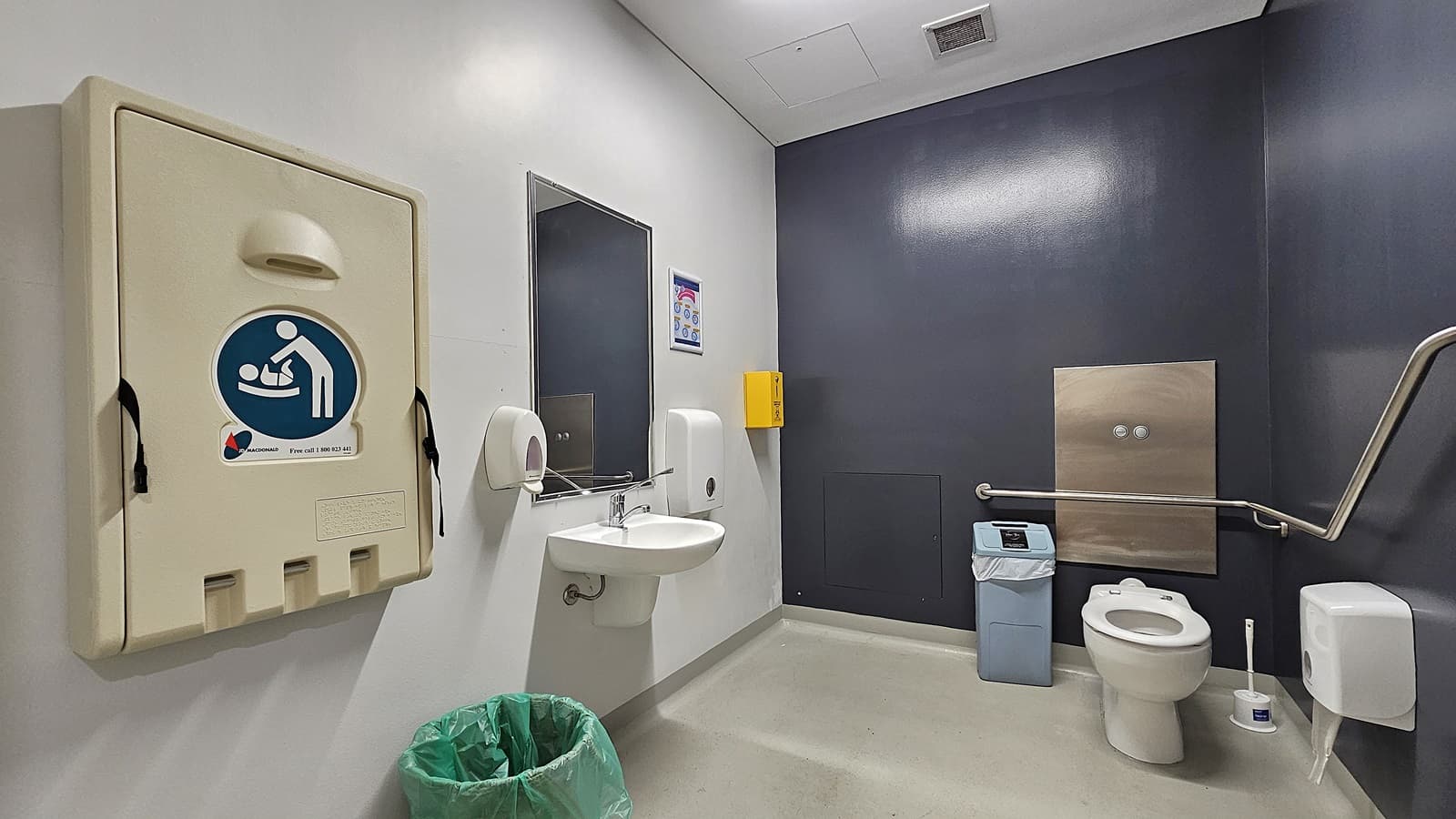 On the left, there is a beige baby changing station mounted on the wall, a green bin on the floor, a sink with a mirror above it, and a soap dispenser. On the right, there is a white toilet with a grey grab rail next to it, a blue bin, and a toilet paper dispenser. In the middle, there is a yellow wall-mounted box. The floor is light grey, and the ceiling is white with recessed lighting and a ventilation panel.