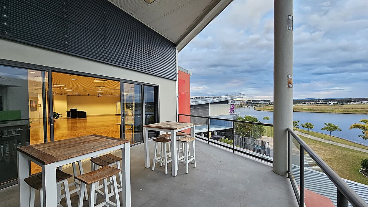 On the left, there is a glass door leading to an indoor area with a wooden floor and ceiling lights. On the right, there is a view of a balcony with a metal railing, overlooking a grassy area with trees and a body of water. In the foreground, there are two white high tables with wooden tops and matching stools. The floor of the balcony is grey concrete, and the ceiling above the balcony is white with recessed lighting.