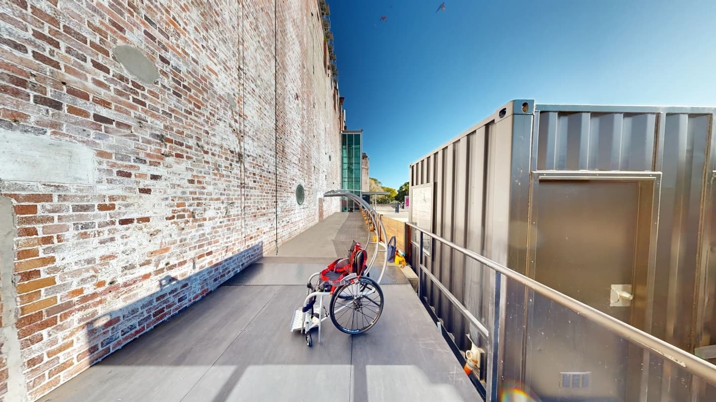 On the left, there is a tall brick wall with circular windows and weathered patches. In the middle, there is a wheelchair with a red harness on a black platform, next to a ramp with a yellow tactile paving strip. In the background, there is a modern glass building and trees. On the right, there is a large grey metal container with a door and a railing along the edge of the ramp. The floor is smooth concrete.