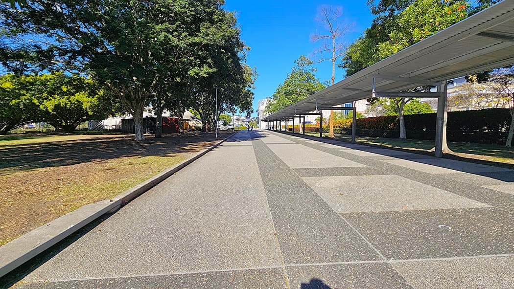 On the left, there is a paved footpath with bollards and a grassy area with trees providing shade. In the middle, there is a road with a "Give Way" sign and a vehicle parked near the bollards. In the background, there are more trees, a few buildings, and a distant view of a park. On the right, there is another road with a large tree casting shadows on the pavement. The floor is a mix of asphalt and concrete.