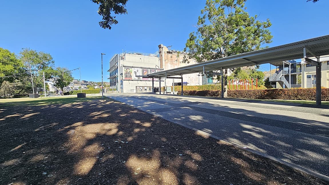 On the left, there is a grassy area with trees casting shadows and a black square planter. In the middle, there is a wide, paved pathway leading towards a multi-storey building with a mural. In the background, there are more buildings and trees. On the right, there is a covered walkway with a metal roof supported by columns, running parallel to the pathway. The floor is a mix of concrete and asphalt.