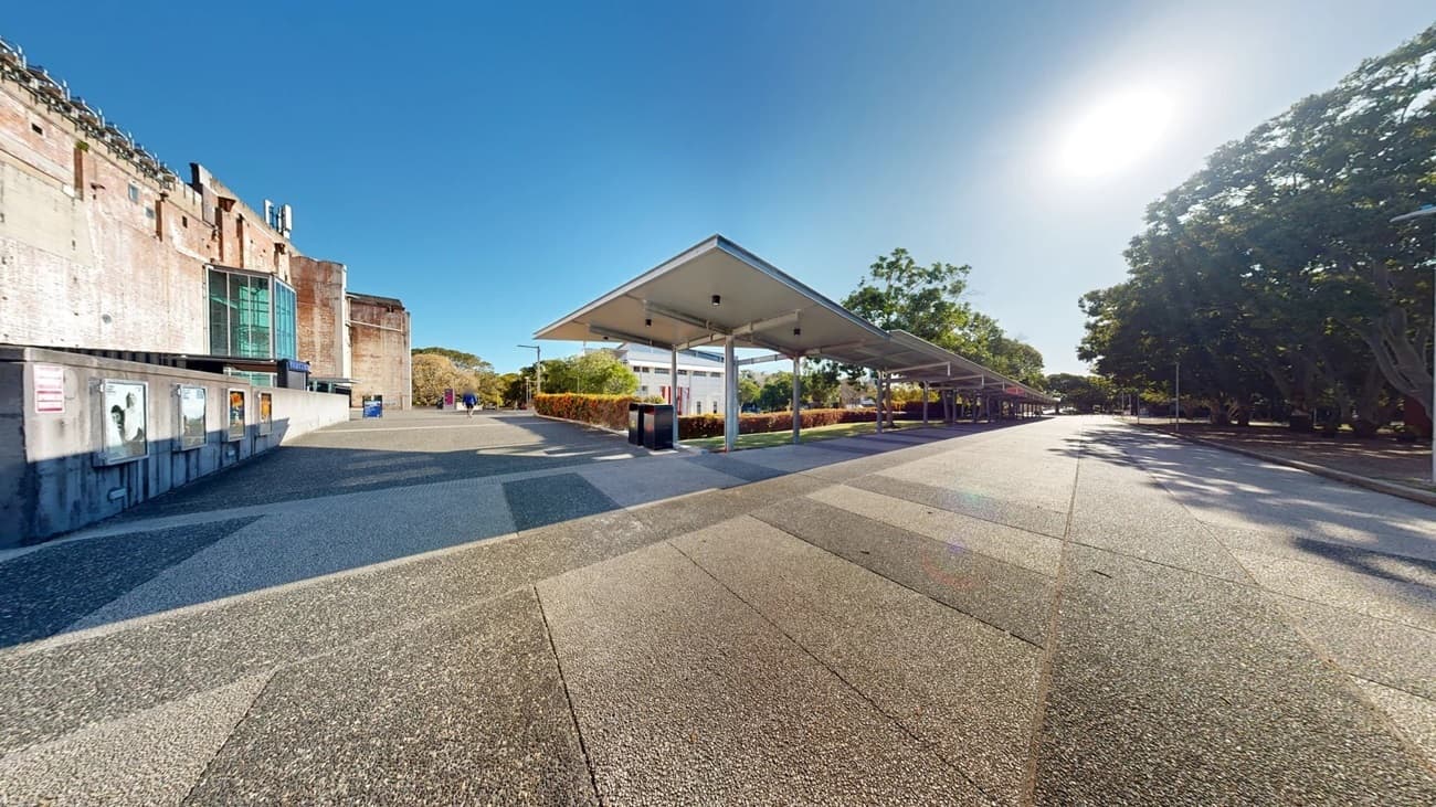 On the left, there is a brick building with large windows and a staircase leading up to it. In the middle, there is a wide, paved area with a mix of light and dark grey tiles, leading to a covered walkway with a metal roof supported by columns. In the background, there are more buildings and trees. On the right, there is a row of trees casting shadows on the pavement. The floor is a mix of light and dark grey concrete.