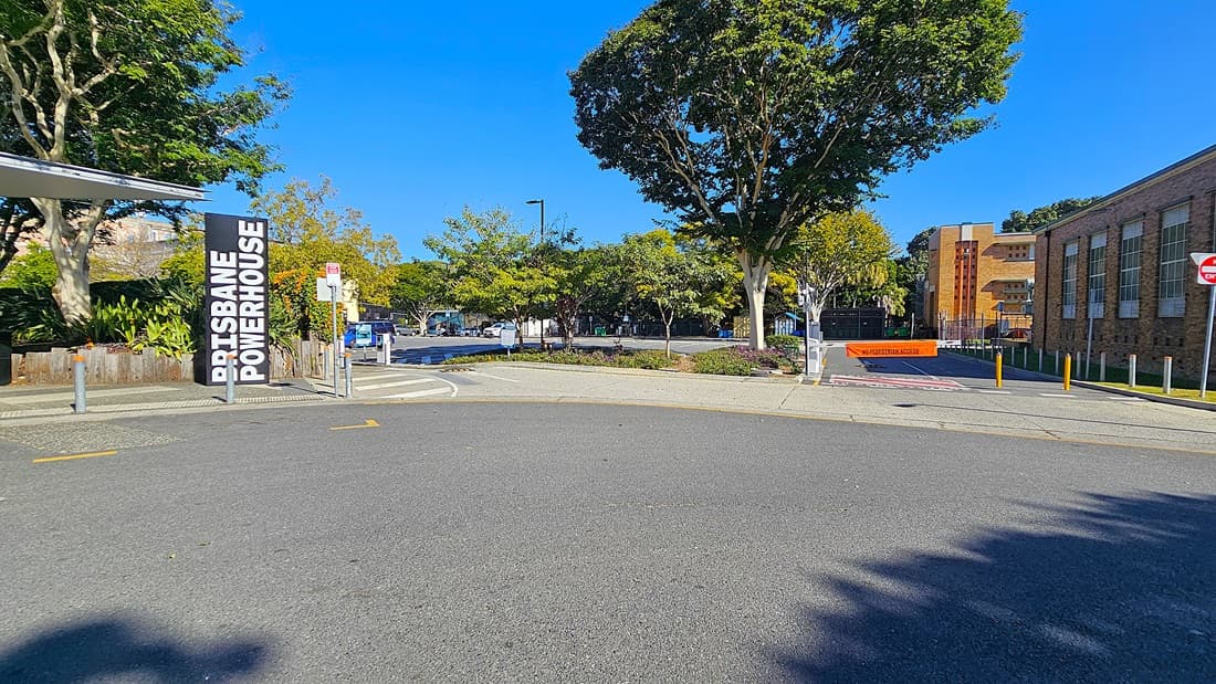 On the left, there is a sign reading "Brisbane Powerhouse" next to a building with a metal roof. In the middle, there is a paved road curving to the right, with a car park and trees in the background. On the right, there is a brick building with large windows and a "No Entry" sign. The floor is asphalt and concrete.