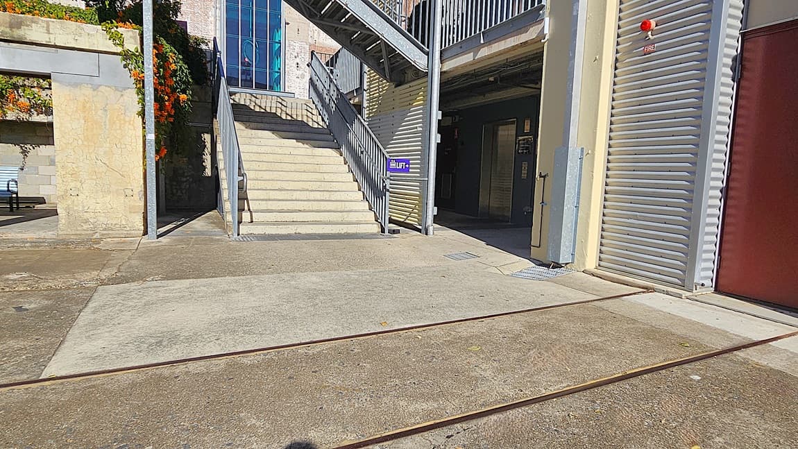 On the left, there is a stone wall with orange flowers growing on it and a metal staircase leading up to a higher level. In the middle, there is a concrete area with metal tracks embedded in the ground, leading to a staircase with a sign reading "Lift" and a door at the top. In the background, there is a building with large windows. On the right, there is a corrugated metal wall with a red door. The floor is concrete with metal tracks.