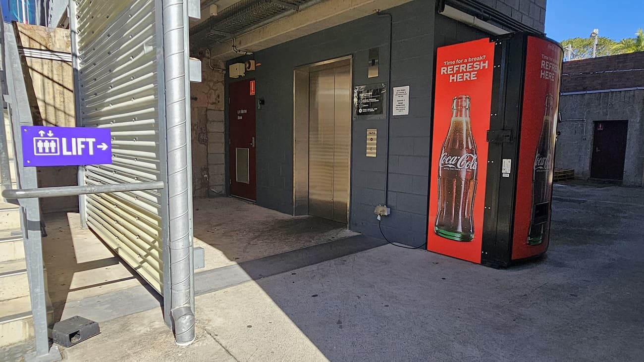 On the left, there is a metal staircase with a purple sign reading "LIFT" and a corrugated metal wall. In the middle, there is a grey building with a lift door and a sign above it. In the foreground, there is a red vending machine with a Coca-Cola advertisement. On the right, there is a concrete wall with a doorway leading to another area. The floor is concrete, and the ceiling is metal with exposed pipes and beams.