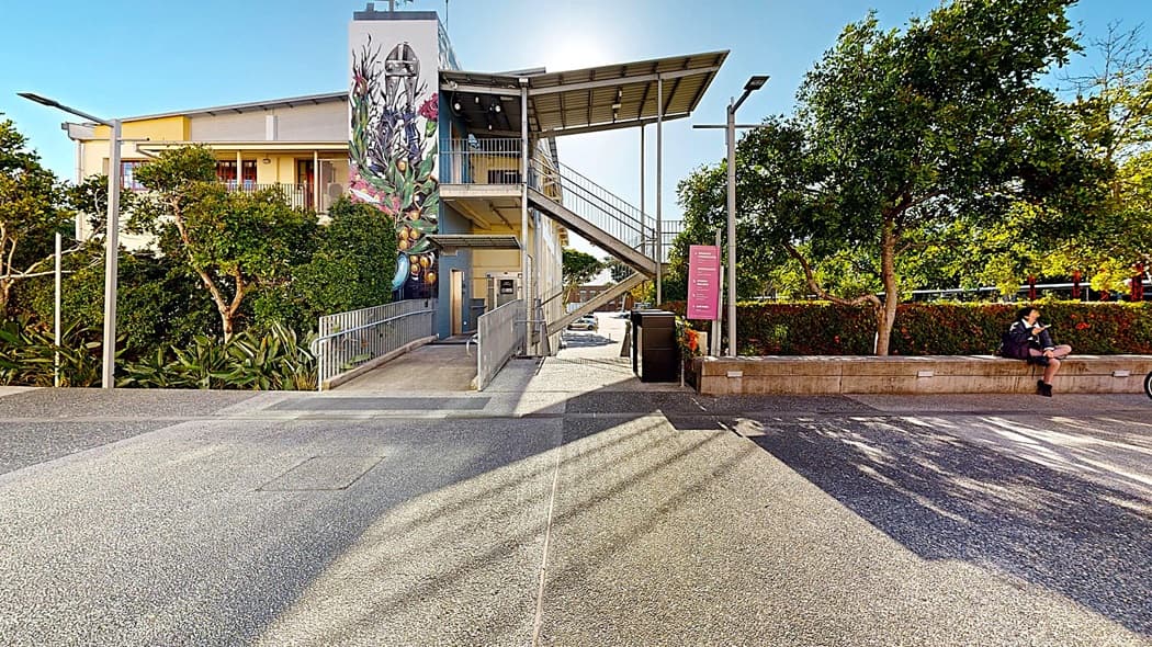 On the left, there is a building with a mural of flowers and a staircase leading up to a covered walkway. In the middle, there is a ramp and a pathway leading to the building's entrance. In the background, there are trees and more parts of the building. On the right, there is a stone bench with two people sitting on it, next to a hedge. The floor is a mix of concrete and asphalt, and the ceiling of the walkway is metal.