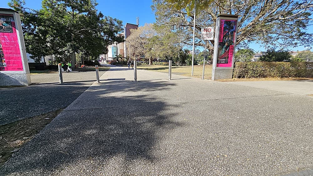 On the left, there are two white cars parked under the shade of large trees. On the right, a blue car is parked next to a white car, with tree branches overhead. In the middle, a paved road leads to a pedestrian area with two tall signs and a few bollards. The background includes more trees, a grassy area, and a building partially visible through the foliage.