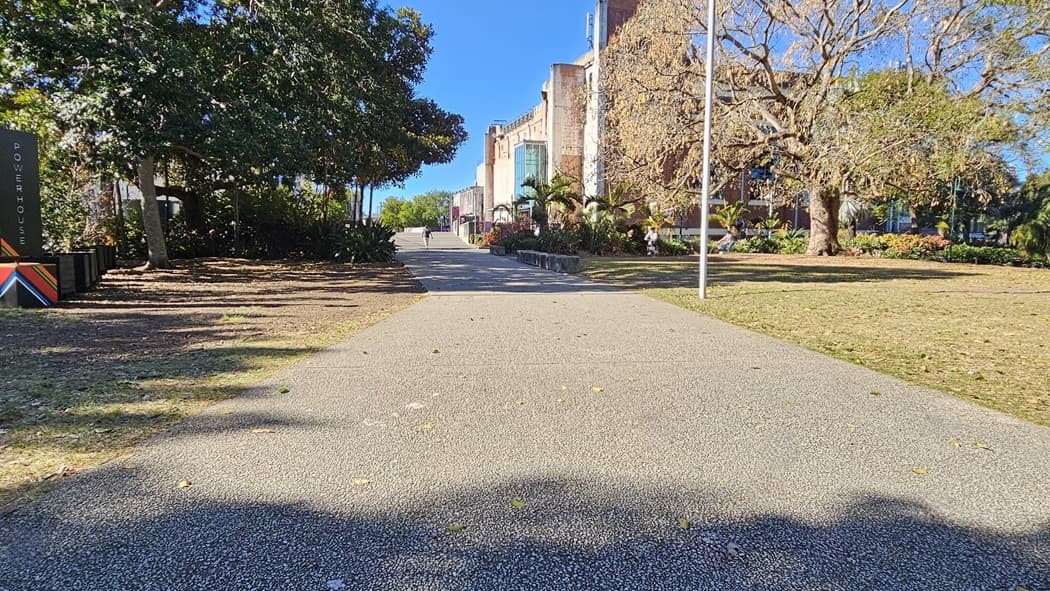 On the left, there are trees casting shadows over a garden bed and a tall sign with text. On the right, a large tree with bare branches stands next to a grassy area. In the middle, a wide, textured pathway leads towards a distant building with multiple windows and a flagpole. The background includes more trees and the building, partially obscured by foliage. The floor is a textured pavement.
