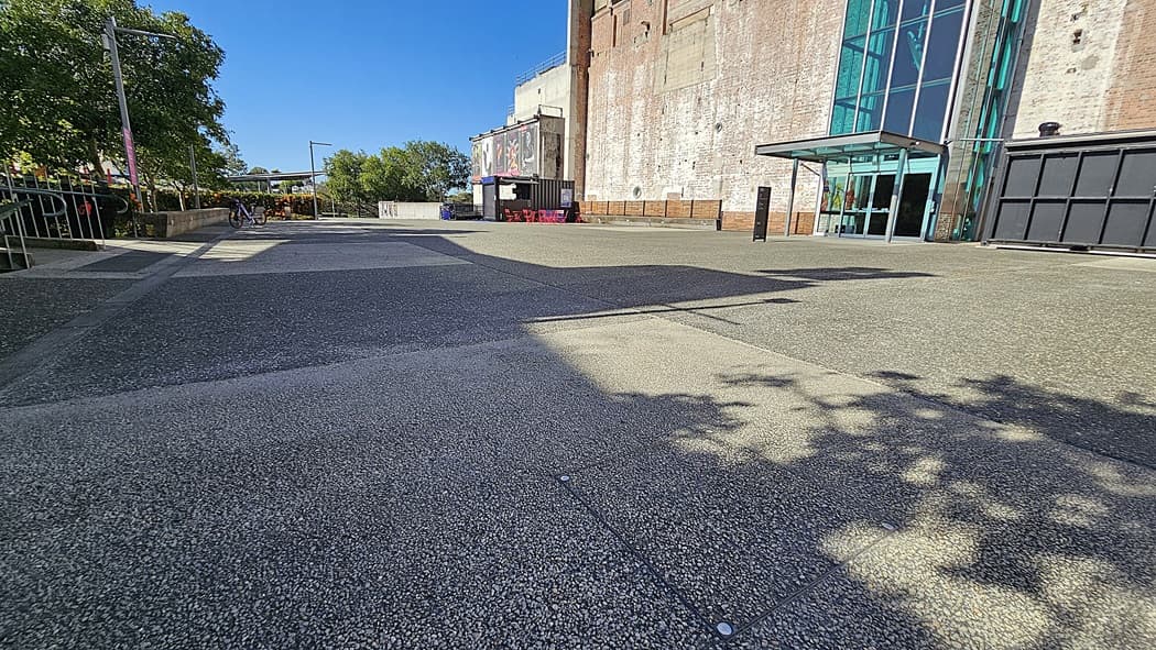 On the left, there are trees casting shadows over a textured pavement and a metal railing. On the right, there is a large brick building with tall windows and a glass entrance. In the middle, a wide, textured pavement leads towards the building. The background includes more trees, a small structure with a canopy, and part of the building. The floor is a textured pavement.