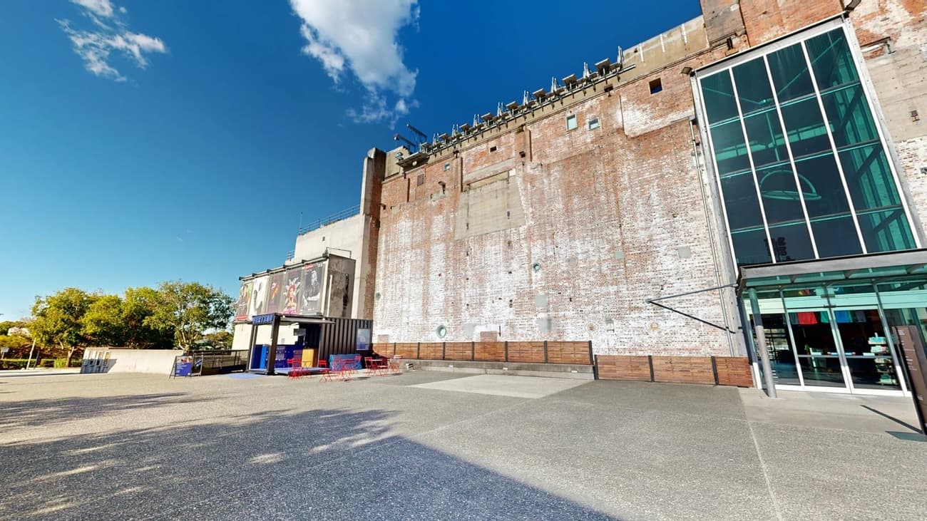 On the left, there are trees and a small structure with blue and red outdoor seating. In the middle, there is a large open concrete area leading to a tall brick wall with weathered paint. On the right, there is a modern glass entrance with a metal canopy and a reflection of the surroundings. The floor is a mix of smooth and textured concrete.