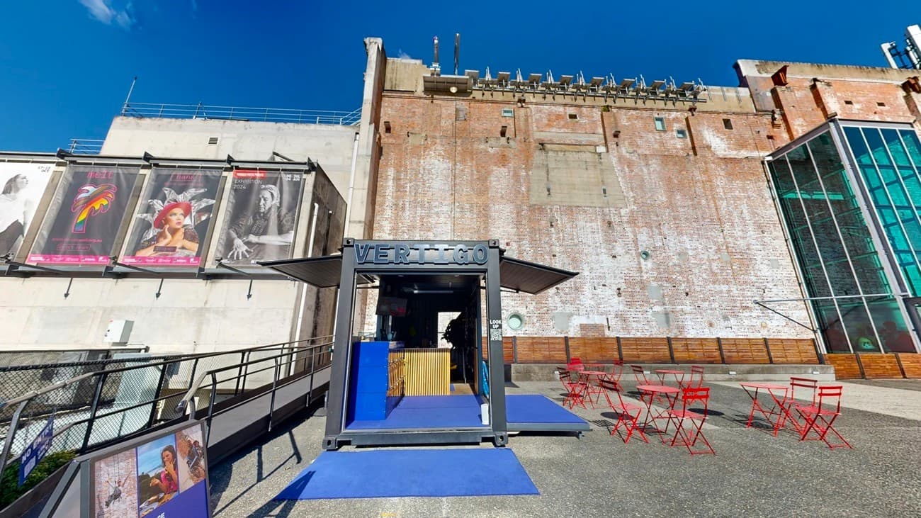 On the left, there is a black metal fence with posters of movies on a white wall. In the middle, there is a black structure with a sign reading "VERTIGO" and blue ramps leading up to it, with a wooden counter inside. In the background, there is a tall, weathered brick wall with various fixtures and a glass section on the right. On the right, there are red metal tables and chairs on a textured concrete floor. The floor is a mix of smooth and textured concrete.