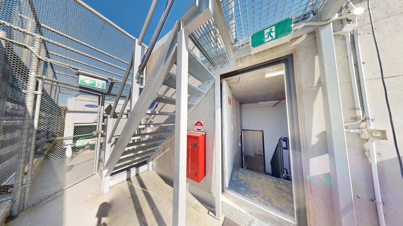 On the left, there is a beige concrete wall with a blue sign reading "NO ENTRY WITHOUT INSTRUCTOR" and metal pipes running vertically. In the middle, there is a metal staircase with yellow steps and a handrail, leading up to a platform. In the background, there are trees and a distant building. On the right, there is a metal railing with horizontal bars and a shadow cast on the floor. The floor is light grey concrete.