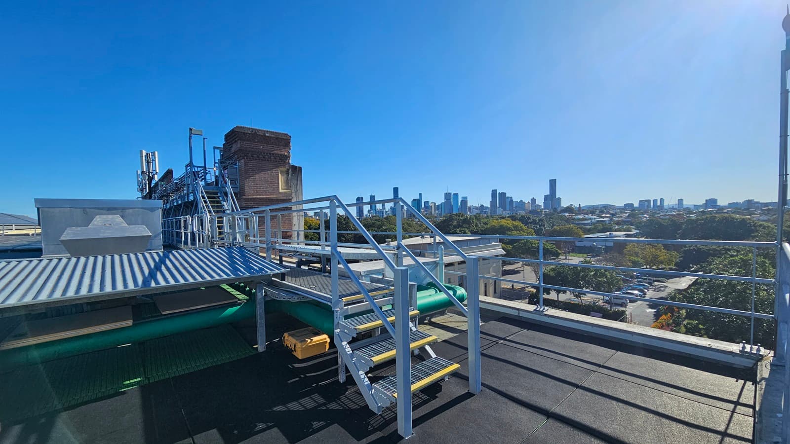 On the left, there is a metal staircase with yellow steps and a handrail, leading up to a platform with a corrugated metal roof. Below the staircase, there are green pipes and a small yellow ladder. In the middle, there is a metal railing and a view of a distant city skyline with tall buildings. On the right, there is a continuation of the metal railing and a view of trees and smaller buildings. The floor is black and appears to be a flat rooftop surface.