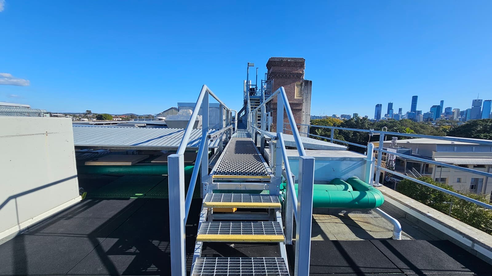 On the left, there is a metal staircase with yellow steps and a handrail, leading up to a platform with a corrugated metal roof. Below the staircase, there are green pipes and a small yellow ladder. In the middle, there is a metal railing and a view of a distant city skyline with tall buildings. On the right, there is a continuation of the metal railing and a view of trees and smaller buildings. The floor is black and appears to be a flat rooftop surface.