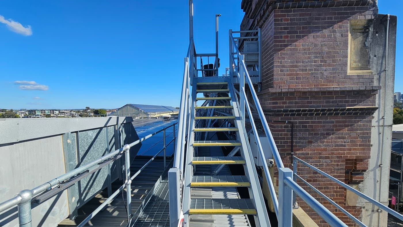 On the left, there is a white building wall with a metal railing extending from it. In the middle, there is a metal staircase with yellow-edged steps and a handrail, leading up to a platform with a corrugated metal roof. Below the platform, there are green pipes. In the background, there is a tall brick structure and a distant city skyline with tall buildings. On the right, there is a continuation of the metal railing and a view of trees and smaller buildings.