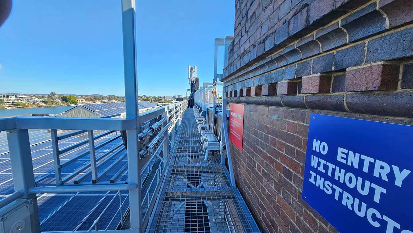 On the left, there is a metal walkway with a railing, running alongside a series of solar panels. In the middle, the walkway continues with metal grating underfoot. In the background, there are distant buildings and a body of water. On the right, there is a brick wall with a blue sign reading "NO ENTRY WITHOUT INSTRUCTOR" and a red sign further along.