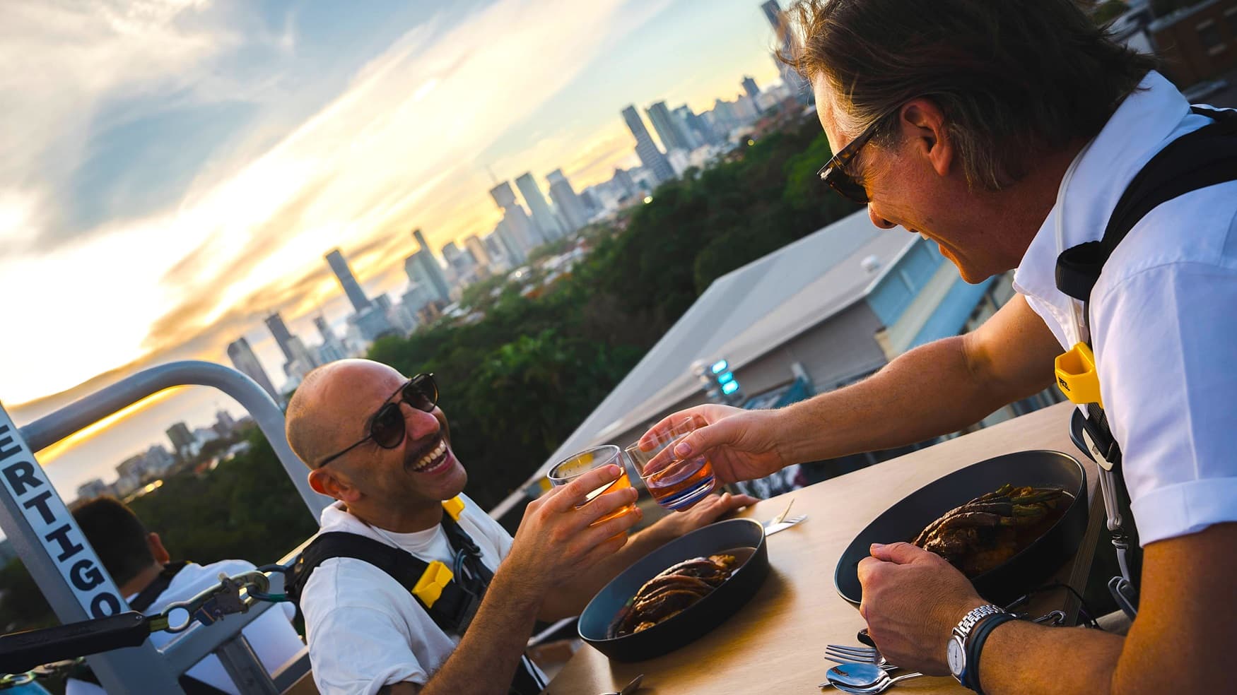 On the left, there is a man wearing sunglasses and a white shirt, holding a drink and smiling. In the middle, there is a wooden table with two plates of food and two drinks. In the background, there is a city skyline with tall buildings and a green area with trees. On the right, there is another man wearing glasses and a white shirt, also holding a drink and smiling.