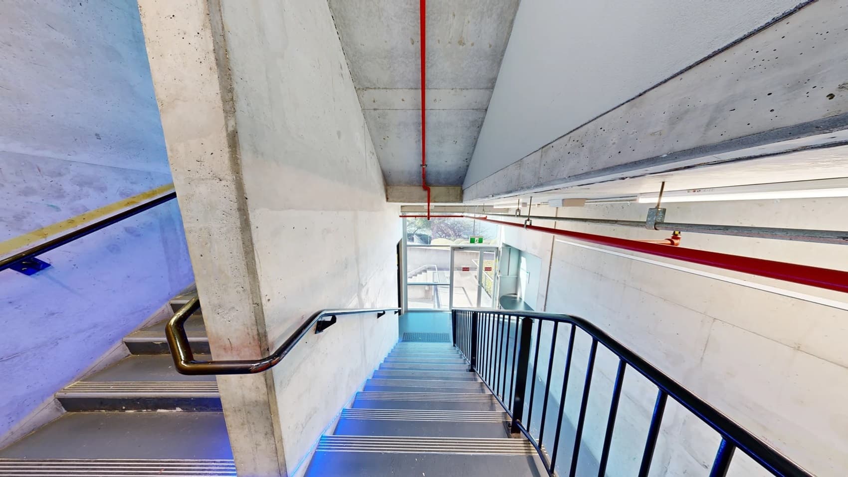 On the left, there is a concrete wall with a black handrail running down a staircase. In the middle, the staircase has metal steps with blue lighting along the edges, leading down to a glass door at the bottom. In the background, through the glass door, there is an exit sign and a glimpse of the outside area. On the right, there is another wall with exposed pipes and electrical conduits on the ceiling. The floor is made of metal steps, and the ceiling is concrete with red and white pipes.