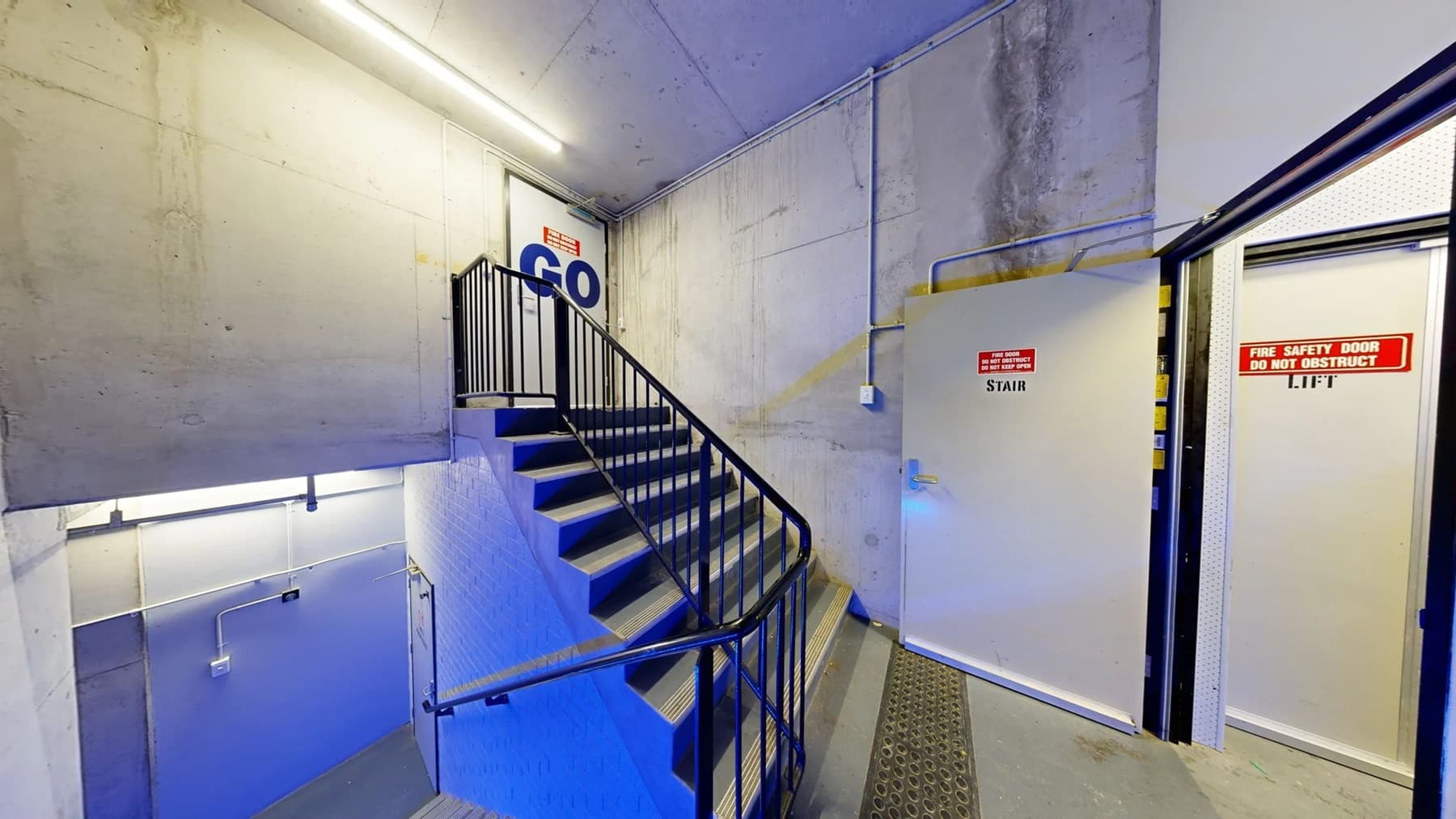 On the left, there is a concrete wall with a black handrail along a staircase. The staircase has black steps with blue lighting underneath. In the middle, the stairs lead up to a white door with a "GO" sign and an emergency exit sign above it. On the right, there is a grey door marked "STAIR" and an open door with a sign reading "FIRE SAFETY DOOR DO NOT OBSTRUCT LIFT." The floor is grey with a metal grate, and the ceiling is concrete with fluorescent lighting.