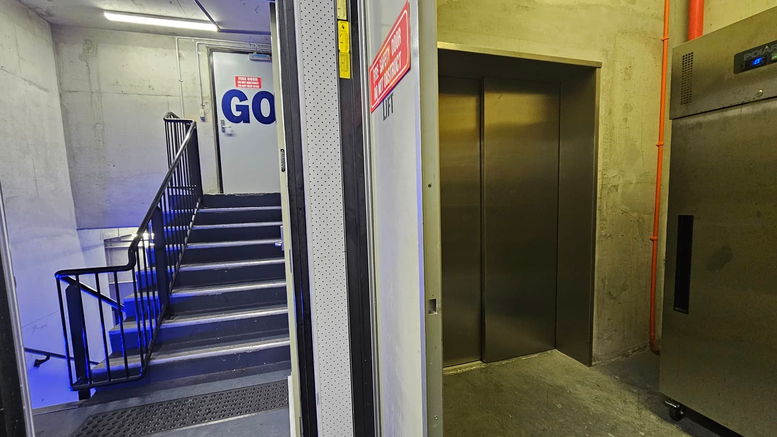On the left, there is a staircase with black steps and blue lighting underneath, leading up to a white door with a "GO" sign and an emergency exit sign above it. The staircase has black railings and a grey mat at the bottom. In the middle, there is a grey wall. On the right, there is a stainless steel lift with closed doors and a control panel, next to a grey wall with exposed orange piping. The floor is grey concrete, and the ceiling is concrete with fluorescent lighting.