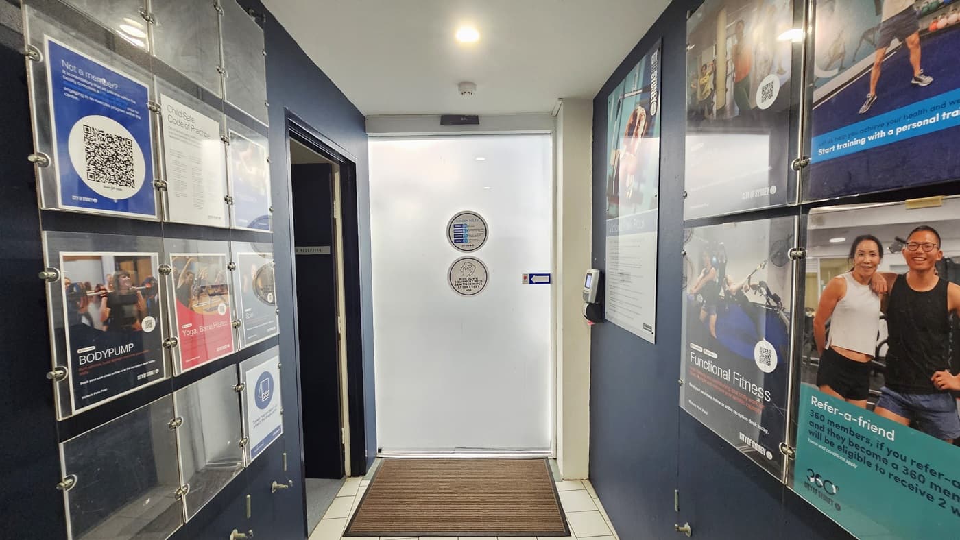 The black wall is equipped with posters and directional signages. To the left is a white sliding door equipped with a swiping machine for badge entry. A brown mat is installed on the doorstep. To the right is a huge retractable banner. The floor is grey, and the ceiling and wall are white with lights.