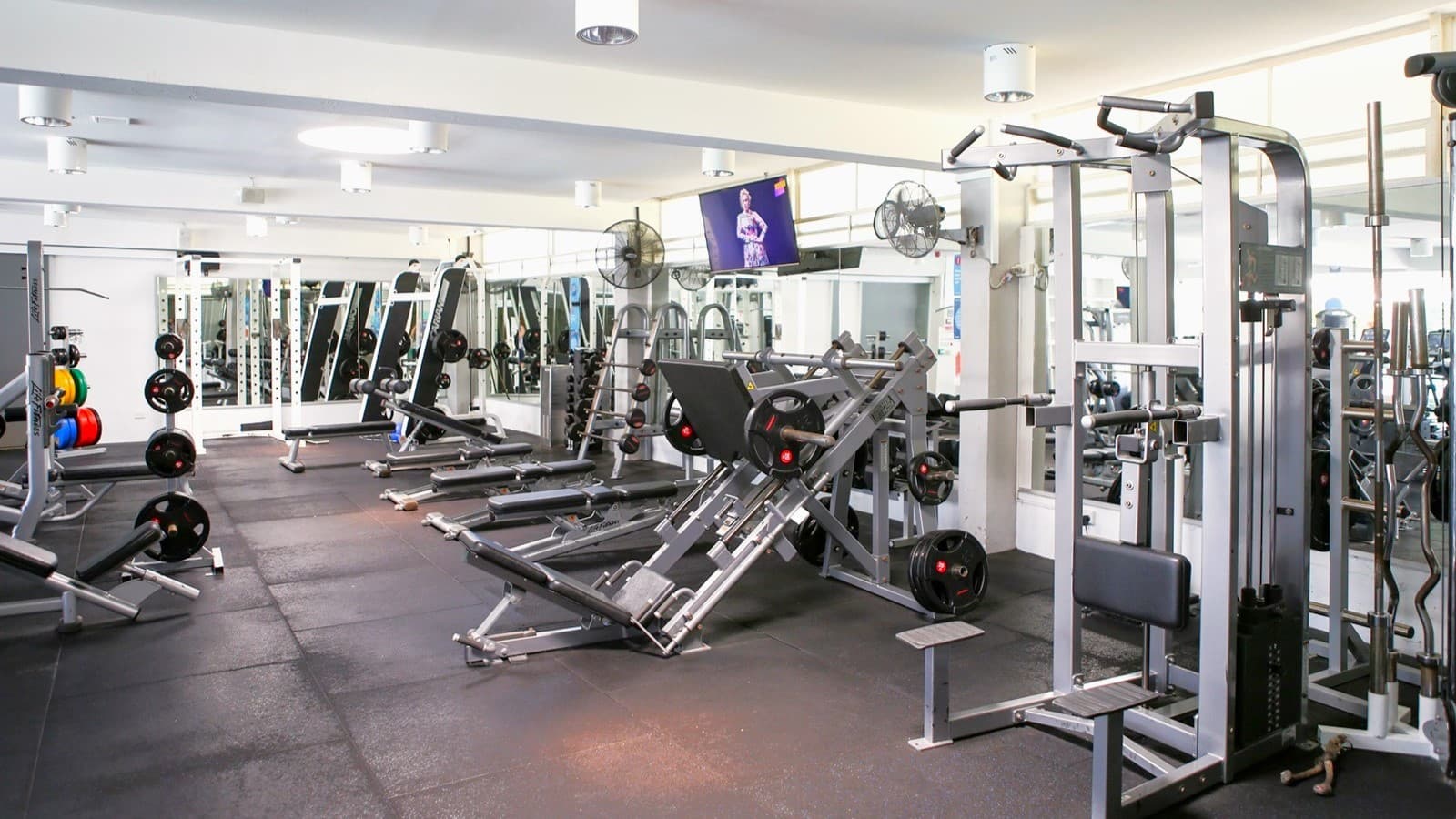 Inside the gym, Lat Pulldown machines, Smith Machines, bench press, and inclined bench press machines are arranged on the black rubber-tiled floor. A flat-screen TV is mounted on the upper right wall along with wall fans. The ceiling is all white with multiple pin lights.