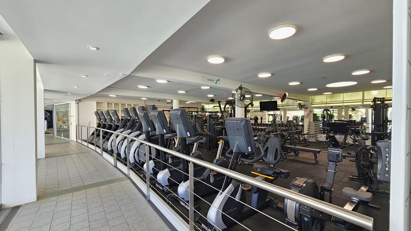 Cardio machines are arranged near the stainless railings. Inside, Lat Pulldown machines, Smith Machines, bench press, and inclined bench press machines, along with other machines, are arranged on the black rubber-tiled floor. A flat-screen TV and wall fans are affixed to the upper wall. To the left is a corridor with grey tiles. The ceiling is white with circular LED lights.