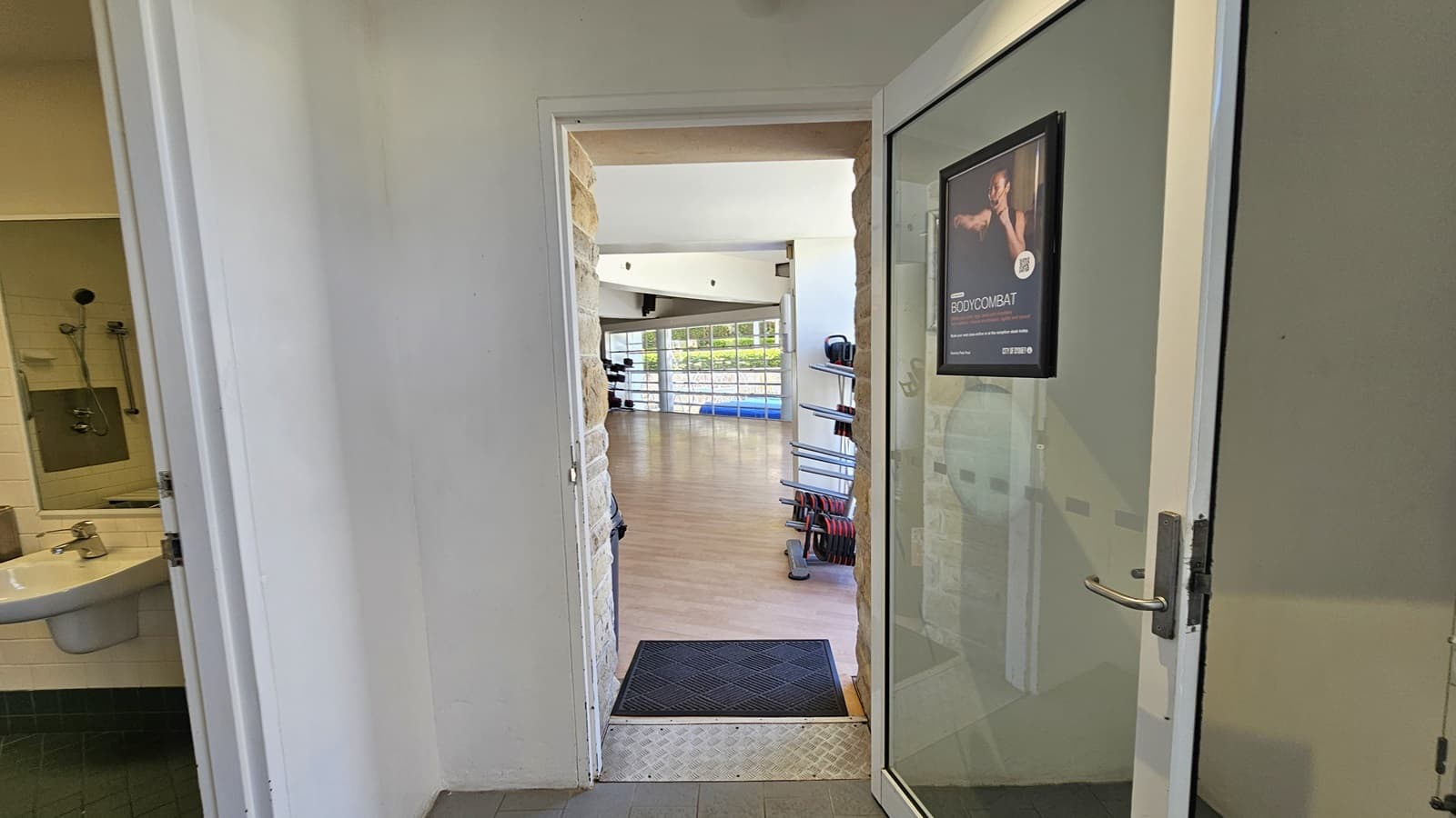 To the left is the bathroom, and adjacent to it is the Exercise Studio, accessible through a manual glass door equipped with a poster. An aluminum step-free ramp is installed on the doorstep along with a non-slippery mat. The studio features a timber floor, and inside, exercise machines are visible.