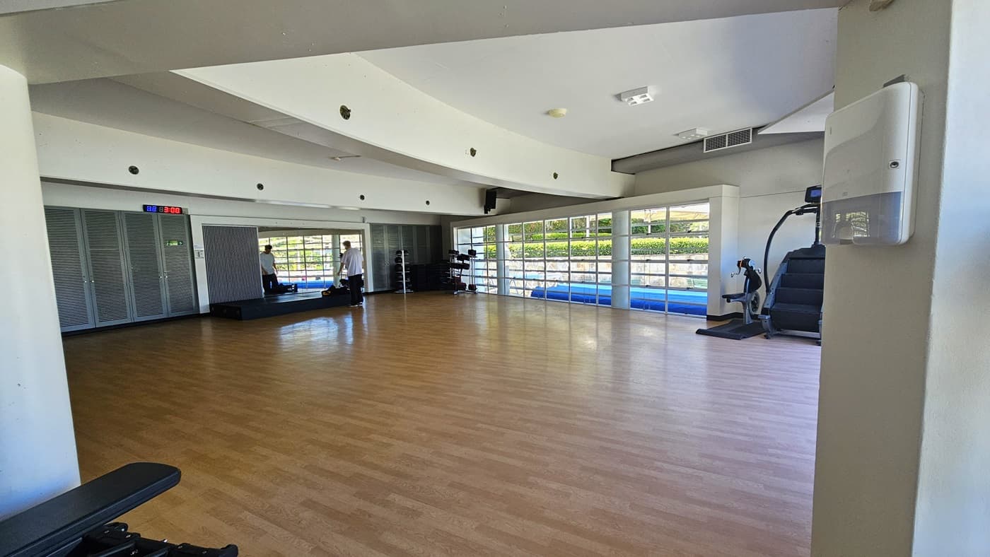 The exercise studio is a spacious area featuring a timber floor. Spin machines and a stair mill are on the right. Weights are arranged in the corner, and a platform is positioned against the mirror. A digital clock is affixed to the upper-left wall. The white ceiling features a cove design with LED lights.