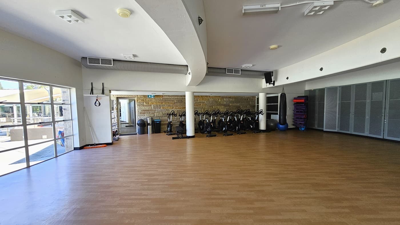 The exercise studio is a spacious area featuring a timber floor. The middle wall has brown bricks, and spin machines are arranged against it. Rubbish bins are placed near the door. In the right corner, there is a punching bag, rolled mats, and stability balls. The white ceiling features a cove design with LED lights. The left wall is made of glass.