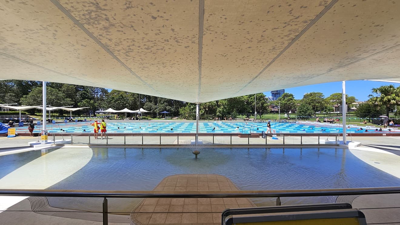 Access to the Outdoor Aquatic Facility is through a manual glass door. Immediately inside, free-standing banners and an aquatic wheelchair are positioned in the middle. Shade cloths overhead provide shade to the Kid's Splash Pool. An aluminum railing separates the upper pool deck and the pool area. Further behind is the lap pool, with pool guards and guests visible.